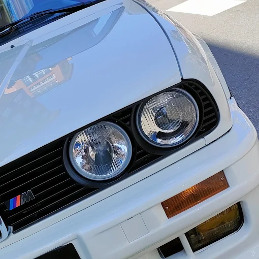 BMW E30 blanco clásico con faros redondos y emblema M tras el detallado de carrocería