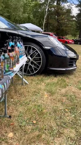Porsche 911 Turbo S negro junto a una mesa de productos Auto Graph Detailing en un evento de coches